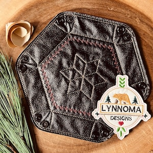Könnte beinhalten: Ein braunes Ledertablett mit geometrischem Muster und einem weißen gestickten Aufnäher mit der Aufschrift "LYNROMA DESIGNS" mit einem Bären und einem Herzen.