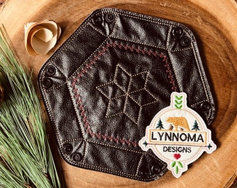Könnte beinhalten: Ein braunes Ledertablett mit geometrischem Muster und einem weißen gestickten Aufnäher mit der Aufschrift "LYNROMA DESIGNS" mit einem Bären und einem Herzen.