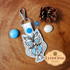 May include: A white owl keychain with blue accents. The owl has a black and white patterned design with a blue heart in the center. The keychain is attached to a silver key ring. The keychain is on a wooden surface with a pine cone and a small blue flower.