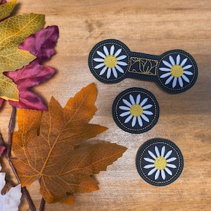 May include: An overhead shot features three daisy-themed patches and a barrette on a wooden surface, alongside colorful autumn leaves. The patches and barrette are black with white petals and yellow centers. The leaves are in shades of yellow, orange, and red.