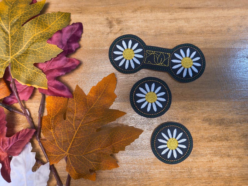 May include: An overhead shot features three daisy-themed patches and a barrette on a wooden surface, alongside colorful autumn leaves. The patches and barrette are black with white petals and yellow centers. The leaves are in shades of yellow, orange, and red.
