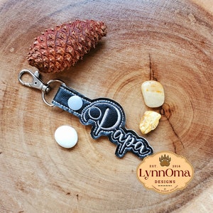 May include: Black leather keychain with a white button and the word "Papa" embroidered in white thread. The keychain is on a wooden surface with a pine cone, two small yellow stones, and a logo for Lynn Oma Designs.
