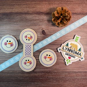 May include: A collection of winter-themed craft supplies on a wooden surface. Includes snowman-faced felt appliques, a light blue snowflake ribbon, a pine cone, and a patch with the text "LYNNOMA DESIGNS" and a bear graphic.