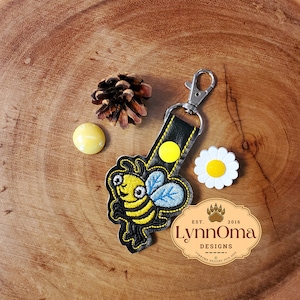 Könnte beinhalten: Ein schwarzer Leder-Schlüsselanhänger mit einem gelben und schwarzen gestickten Bienen-Anhänger. Der Schlüsselanhänger hat einen silbernen Metallverschluss und einen gelben Knopf. Der Text "LynnOma Designs Est. 2016" befindet sich auf einem braunen ovalen Anhänger.