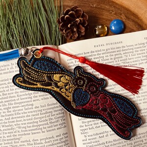 May include: A black leather bookmark with gold and red embroidery depicting a bird with outstretched wings. The bookmark has a red tassel and a blue tassel attached to it.