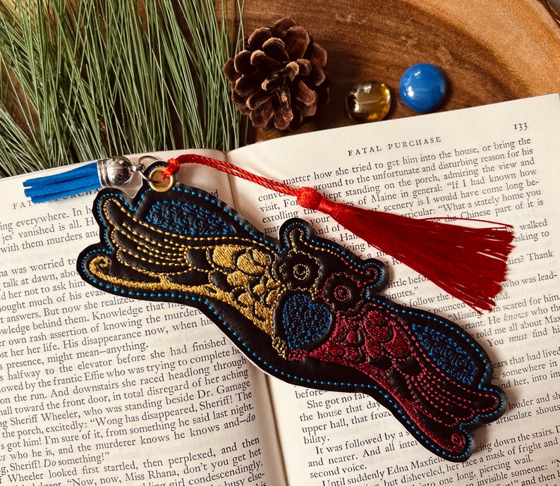 May include: A black leather bookmark with gold and red embroidery depicting a bird with outstretched wings. The bookmark has a red tassel and a blue tassel attached to it.