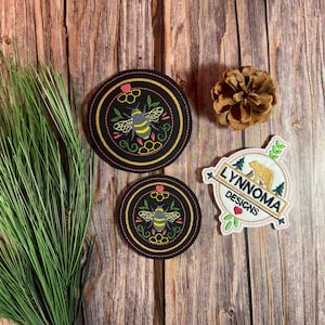 May include: Two black circular embroidered patches with a bee design, a patch with the text "LYNNOMA DESIGNS" and a pinecone on a wooden surface. The bee patches have yellow, pink, and green accents.