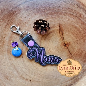 May include: A black leather keychain with a purple embroidered name "Nana" and a pink circle. The keychain is attached to a silver split ring. The keychain is on a wooden surface with a small pine cone.