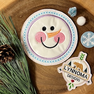 May include: A white snowman face with pink cheeks and an orange nose, embroidered on a round blue and white fabric patch. The patch is surrounded by a purple border with white stitching. The patch is labeled "Lynnnoma Designs" in a brown and white font.