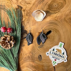 May include: A pair of black and purple leaf-shaped earrings with silver hooks. The earrings are on a wooden surface with a small pine branch and a white seashell.