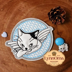 Peut inclure: Patch brodée thermocollante représentant un chat blanc avec des marques noires, portant des ailes d'ange et dormant sur une lune bleue et blanche. Le patch est entouré d'un contour bleu et porte le texte "LynnOma Designs" et "Est. 2016" en dessous.