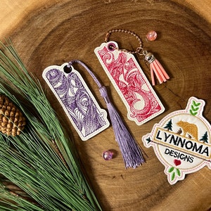 Puede incluir: Dos marcapáginas bordados en morado y rojo con borlas, y un parche bordado blanco con un oso y el texto "Lynnnoma Designs".