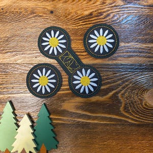 May include: Embroidered daisy patches in black, white, and yellow, arranged on a wooden surface. The patches include three circular designs and one elongated design. Three small wooden tree cutouts are also present.