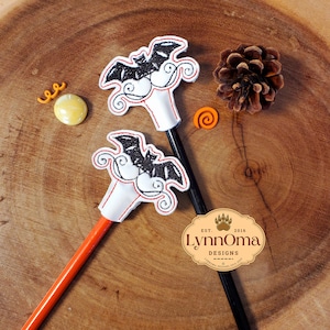 May include: Two black and white bat pencil toppers with red stitching on a wooden surface. The pencil toppers are attached to pencils, one black and one orange. The pencils are on a wooden surface with a pine cone and a small orange swirl.