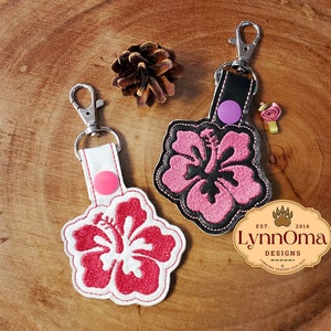 May include: Two hibiscus flower keychains with silver clasps. One keychain has a white strap and a red flower design, the other has a black strap and a pink flower design. A pinecone and a logo are also visible.