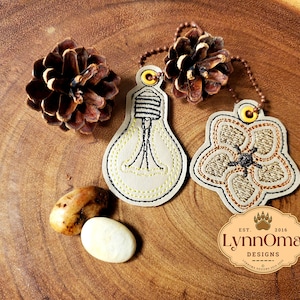 May include: Two embroidered fabric keychains, one shaped like a light bulb and the other like a flower. The light bulb keychain is beige with yellow stitching and the flower keychain is beige with brown stitching. The keychains are on a wooden surface with a pine cone and two small stones.