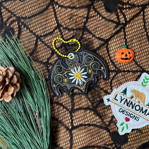 May include: Black and white embroidered bat with a yellow daisy in the center. The bat is attached to a silver chain and is hanging on a black spiderweb-patterned fabric. The fabric is also decorated with a small orange pumpkin and a white embroidered patch with the text "Lynnnoma Designs" and a brown bear.