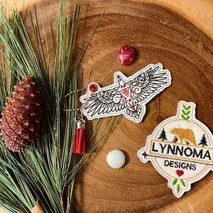 May include: Two embroidered patches on a wooden surface. The first patch features a stylized black and white bird with red accents. The second patch is round and features a brown bear with the text "Lynnnoma Designs".