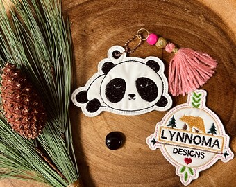 May include: A white panda bear with black eyes and ears, lying down on a white fabric with a pink tassel keychain attached. The keychain has a gold bead and a pink bead. The panda is part of a set of embroidered patches with the text "LYNNOMA DESIGNS" on a white fabric with a brown bear and green trees.