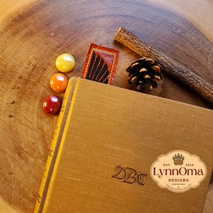 May include: A brown leather bookmark with a black and red feather design. The bookmark is on a wooden surface with a book, a pine cone, and a twig.