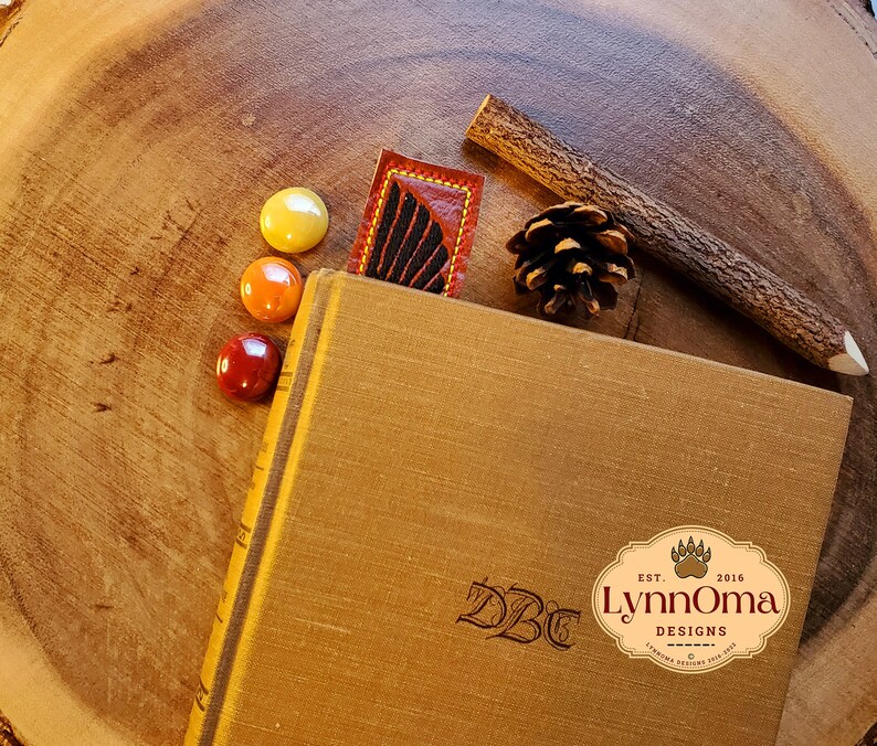 May include: A brown leather bookmark with a black and red feather design. The bookmark is on a wooden surface with a book, a pine cone, and a twig.