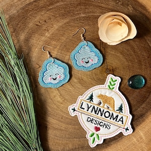 May include: A pair of blue and white cloud-shaped earrings with a smiling face. The earrings are hanging from gold hooks. The earrings are on a wooden surface with a green plant and a small wooden flower.