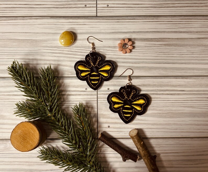 May include: A pair of black and gold bee earrings. The bees are embroidered with yellow thread and have black wings.