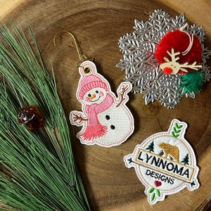 May include: Three embroidered Christmas ornaments: a snowman with a pink scarf, a snowflake, and a brown bear with the text "Lynnnoma Designs".
