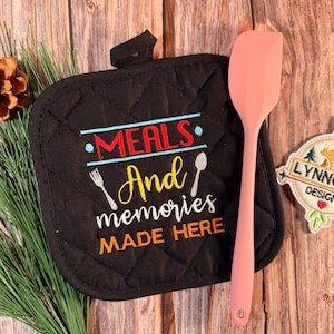 May include: Black quilted pot holder with the text "MEALS And memories MADE HERE" in colorful embroidery. A pink spatula and a "LYNNOMA DESIGNS" patch are next to the pot holder. The background is a wooden surface.