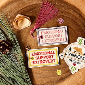 May include: Embroidered patches and tags with the words "EMOTIONAL SUPPORT EXTROVERT" in a variety of colors. A patch with a bear and the text "LYNNOMA DESIGNS" is also visible. The items are arranged on a wooden surface.