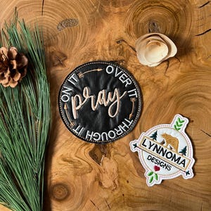 Puede incluir: Dos parches bordados sobre una superficie de madera. El primer parche es negro con costuras blancas y dice "Pray on it, through it, over it". El segundo parche es blanco con costuras doradas y dice "Lynnoma Designs" con un oso y pinos.