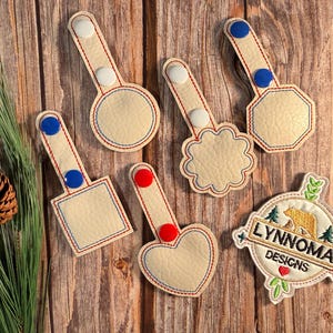 May include: A collection of cream-colored, stitched patches in various shapes: circle, square, heart, and octagon. Each has a handle with button accents. A patch with the text "LYNNOMA DESIGNS" and a bear graphic is also present.