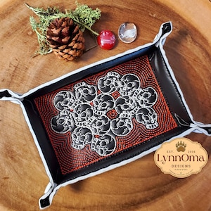 Könnte beinhalten: Schwarzes Leder-Würfeltablett mit einem weißen Totenkopfdesign und roten Nähten. Das Tablett hat einen weißen Rand und liegt auf einer Holzoberfläche. Der Text "LynnOma Designs" ist in der unteren rechten Ecke sichtbar.