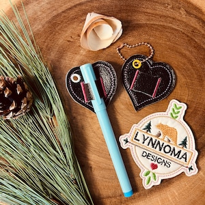 Op de afbeelding: Twee zwarte leren penhouders in de vorm van een hart met roze stiksels en een blauwe pen. Een wit en bruin geborduurd patch met de tekst "LYNNOMA DESIGNS" en een illustratie van een bruine beer.