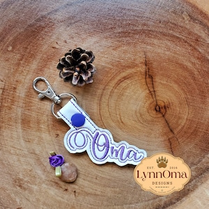Könnte beinhalten: Weißer Stoffschlüsselanhänger mit einem lila bestickten "oma" und einem blauen Knopf. Der Schlüsselanhänger hat einen silbernen Metallring und einen Karabiner. Der Hintergrund ist eine Holzoberfläche.