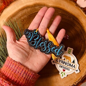 Puede incluir: Un adorno bordado en negro y azul con la palabra "Blessed" con un borla amarilla y un logotipo bordado en blanco, marr&oacute;n y verde que dice "Lynnnoma Designs".