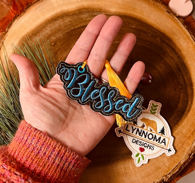 Puede incluir: Un adorno bordado en negro y azul con la palabra "Blessed" con un borla amarilla y un logotipo bordado en blanco, marr&oacute;n y verde que dice "Lynnnoma Designs".