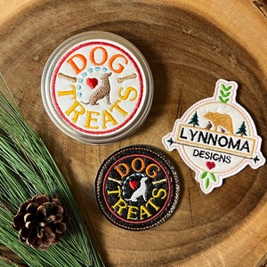 Könnte beinhalten: Drei gestickte Aufnäher auf einer Holzoberfläche. Ein Aufnäher zeigt "DOG TREATS" mit einer Hundeillustration. Ein weiterer Aufnäher hat einen Bären und den Text "LYNNOMA DESIGNS". Der dritte Aufnäher befindet sich in einer Metalldose und zeigt "DOG TREATS".