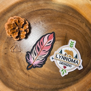 Peut inclure: Plusieurs écussons décoratifs et un cône de pin sur une surface en bois. Les écussons comprennent un motif de plume rose et noir et un écusson circulaire avec un ours et le texte "LYNNOMA DESIGNS".