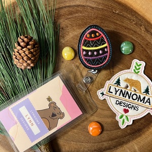 May include: A close-up of a badge holder with a colorful embroidered Easter egg charm, a brown bear logo patch with the text "Lynnnoma Designs", and a card with the text "Lynnnoma Designs".