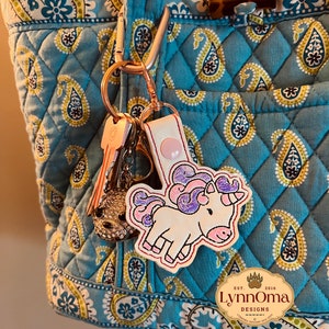 May include: A white and pink unicorn keyring with purple hair and a pink button is attached to a rose gold carabiner with keys. The keyring hangs from a blue paisley patterned tote bag.