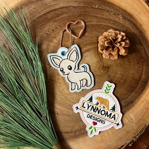 May include: A white chihuahua keychain with blue eyes and a pink heart keychain charm. The keychain is attached to a white circle with a brown bear and green trees. The circle has the text "Lynnoma Designs" in gold lettering.
