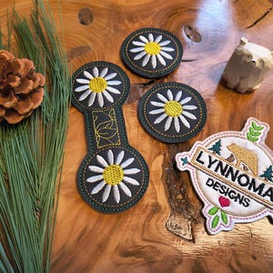 May include: An assortment of embroidered patches and decorative items on a wooden surface. The patches feature daisy designs and a logo with the text "LYNNOMA DESIGNS". A wooden bear figurine and pine needles are also present.