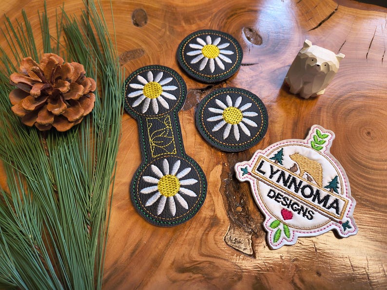 May include: An assortment of embroidered patches and decorative items on a wooden surface. The patches feature daisy designs and a logo with the text "LYNNOMA DESIGNS". A wooden bear figurine and pine needles are also present.