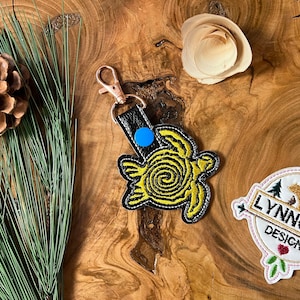 May include: A yellow and black embroidered turtle keychain with a blue accent. The keychain is on a wooden surface with a pine branch and a small wooden flower.
