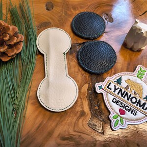 May include: A collection of handmade items on a wooden surface. Includes a beige leather key fob, two black leather coasters, a wooden bear figurine, a pine cone, and a patch with the text "LYNNOMA DESIGNS".