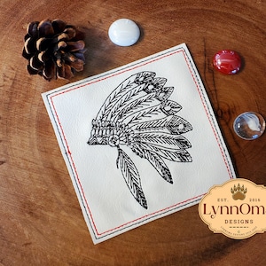 Puede incluir: Un diseño bordado en blanco y negro de un tocado nativo americano sobre un cuadrado de tela blanca con costuras rojas alrededor de los bordes. El cuadrado de tela está sobre una superficie de madera. El diseño es de LynnOma Designs, establecido en 2016.
