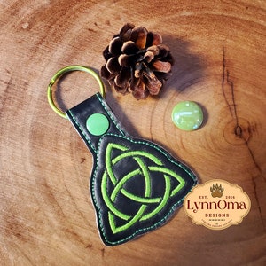May include: A black leather keychain with a green Celtic knot design. The keychain has a gold metal ring and a green button. The keychain is on a wooden surface.