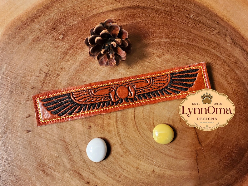 May include: A brown leather bookmark with a black embroidered design of a winged scarab beetle. The bookmark has yellow stitching around the edges and the text "LynnOma Designs Est. 2016" is visible in the lower right corner.