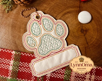 May include: White paw print ornament with a red and green stitched border. The ornament has a blank space for personalization and a gold chain for hanging. The ornament is made by Lynn Oma Designs, established in 2016.